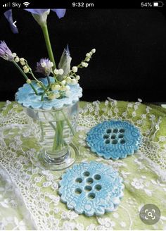 a vase filled with purple flowers sitting on top of a lace covered table cloth next to two blue buttons