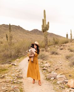 a woman holding a baby in her arms while standing on a dirt path next to a cactus