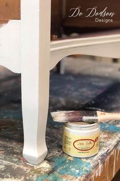 a paint can sitting on top of a table next to a white painted chair with a brush in it