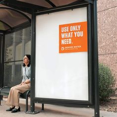 a woman sitting on a bench in front of a bus stop with an orange sign that says use only what you need