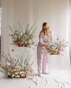 a woman standing next to two flower arrangements on a white backdrop with flowers in them