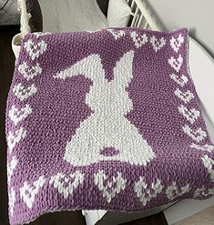 a purple and white knitted pillow sitting on top of a wooden floor next to a chair