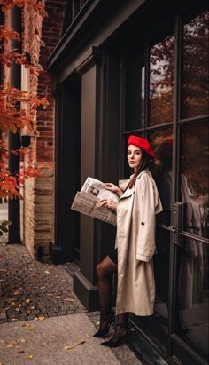 a woman in a red hat and trench coat is holding a newspaper while leaning against a window