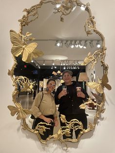 two women taking a selfie in front of a mirror with gold butterflies on it