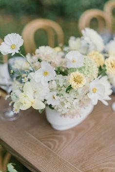 white and yellow flowers are in a vase on a wooden table with chairs around it