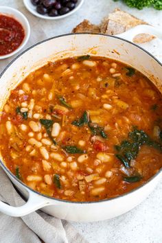 a white pot filled with beans and greens next to bowls of other foods on a table