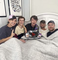 four young men sitting on a bed with one giving the thumbs up and holding something in his hand