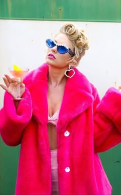 a woman in a pink fur coat and sunglasses holding a drink with both hands while standing next to a green wall