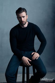 a man sitting on top of a stool in front of a gray wall with his hands on his hips