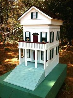 a white doll house sitting on top of a green platform in front of some trees