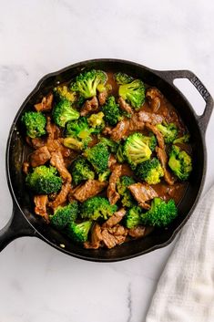 beef and broccoli cooking in a skillet on a white countertop next to a towel