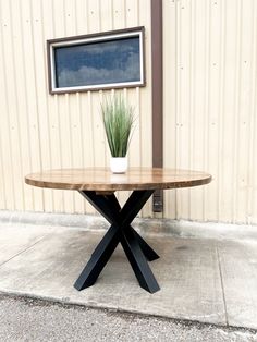 a round wooden table sitting in front of a building with a potted plant on it