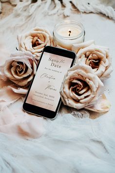 a cell phone sitting on top of a white blanket next to some flowers and a candle