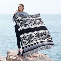 a woman standing on top of a rock next to the ocean holding up a black and white blanket