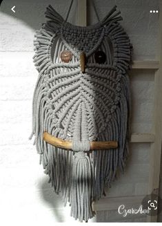 an owl made out of yarn hanging on a wall with a wooden stick in it's mouth