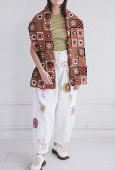 a woman standing in front of a wall wearing white pants and a brown jacket with colorful patches on it