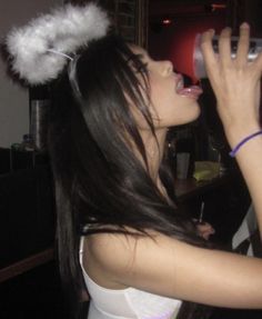 a woman with long black hair is drinking from a cup and wearing a white feather headband