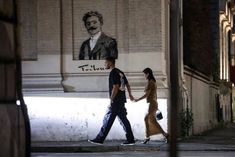 two people walking down the street in front of a wall with a portrait of a man and woman on it