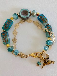 Lovely double strand beaded Premium Czech glass flower bracelet featuring table cut matte aqua with bronze rectangular beads, gold etched ivory/aqua rondelles, fire polished aqua Aurora Borealis crystals, and single 14 mm turquoise Picasso pansy flower. All accented with antique gold plated daisies and shiny gold cubes. Satin Hamilton rose chain adds the perfect delicate second strand. Premium gold electroplated brass chain in satin finish with intricate rose flower pattern. Bracelet is beaded o Gold Hand-strung Czech Glass Bracelets, Gold Czech Glass Hand-strung Bracelet, Hand-strung Gold Czech Glass Jewelry, Hand-strung Gold Jewelry With Czech Glass, Gold Czech Glass Jewelry, Hand-strung, Adjustable Gold Beaded Bracelets Made Of Czech Glass, Adjustable Gold Beaded Bracelets With Czech Glass, Adjustable Gold Beaded Bracelets In Czech Glass, Gold Bohemian Glass Beaded Bracelets