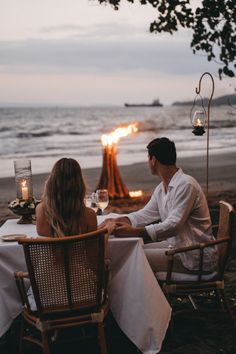 two people sitting at a table with wine glasses in front of an open fire pit on the beach