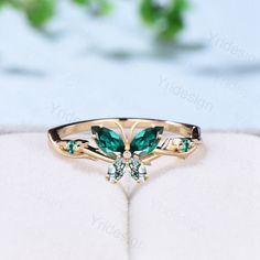 a gold ring with green stones and leaves on it's side, sitting on a white surface