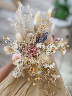 an arrangement of dried flowers is displayed on a table top with someone's hand in the background