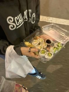 a person holding a plastic container filled with sushi and other food on top of a table