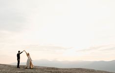 a bride and groom standing on top of a mountain holding hands in the air with mountains in the background