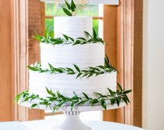 a white wedding cake with greenery on top is sitting on a table in front of a window