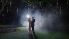 a man and woman standing in the grass at night with lights shining down on them