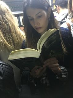 a woman reading a book while sitting on a train seat with other people in the background