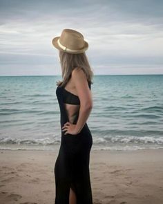 a woman standing on top of a beach next to the ocean wearing a black dress