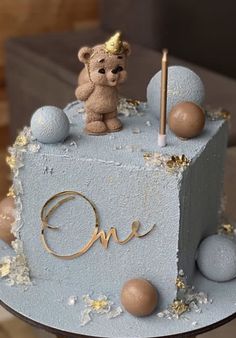 a blue cake with a teddy bear on top and the word one spelled out in gold