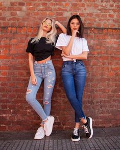 two women standing next to each other in front of a brick wall wearing ripped jeans