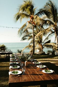 a wooden table topped with white plates covered in food next to palm trees and the ocean