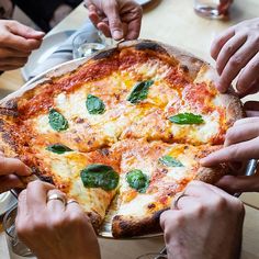 several people are holding their hands over a pizza that is on a table with silverware