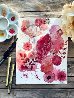 watercolor flowers on paper next to paints and paintbrushes sitting on a wooden table