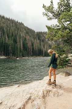 a woman is standing on the edge of a cliff overlooking a body of water with trees in the background