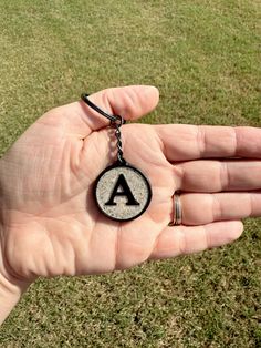 a hand holding a keychain with the letter a on it in front of some grass