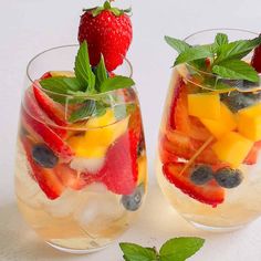 two glasses filled with fruit and ice on top of a white table next to each other