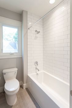 a white toilet sitting next to a bath tub in a bathroom under a window on top of a hard wood floor