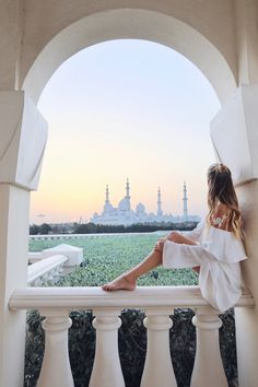 a woman sitting on top of a white balcony next to a lush green field with buildings in the background