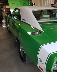 a green and white muscle car parked in a garage next to another one with the hood up