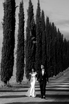 a bride and groom walking down the road in front of tall, slender cypress trees