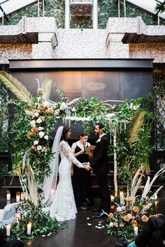 a bride and groom standing in front of an arch with greenery, candles and flowers