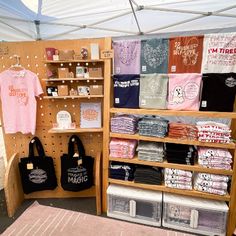 a display with t - shirts and totes on shelves in front of a tent