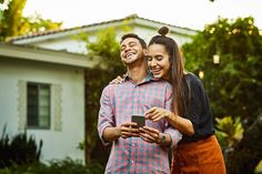 a man and woman standing in front of a house looking at a cell phone together