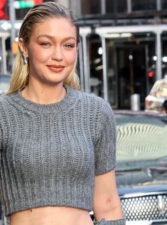 a woman in grey sweater and jeans walking on street next to parked car with other cars behind her