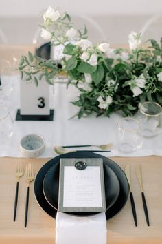 a place setting with black plates, silverware and white flowers in vases on the table