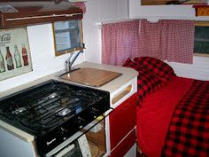 a kitchen with a stove top oven next to a red bed spread and window curtains